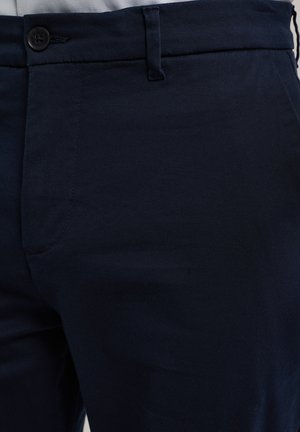 Nahaufnahme von dunkelblauen Hosen mit einem schwarzen Knopf, Gürtelöse und Seitentasche bei einer Person, die ein weißes Hemd trägt.