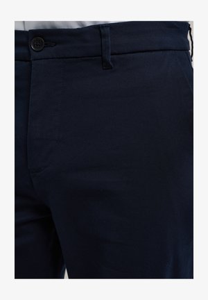 Nahaufnahme von dunkelblauen Hosen mit einem schwarzen Knopf, Gürtelöse und Seitentasche bei einer Person, die ein weißes Hemd trägt.