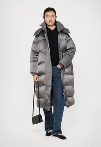 Grijze gewatteerde jas met een hoge kraag, ritssluiting en geribbelde manchetten, gedragen over een gestructureerde zwarte trui en gecombineerd met uitlopende jeans.