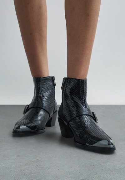 Cizme negre, texturate ca pielea de șarpe, până la glezne, cu capace metalice la vârf și tocuri bloc purtate pe picioare goale, pe un fundal neutru de podea și perete.