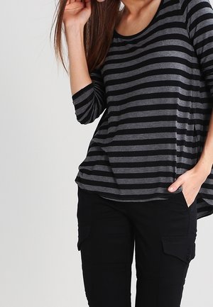 Femme portant un shirt à manches longues rayé noir et gris avec un pantalon cargo noir, une main dans la poche et l'autre touchant ses cheveux.