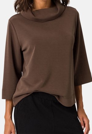 Femme portant un haut marron à manches trois-quarts avec un col roulé ample et un pantalon noir sur un fond uni.