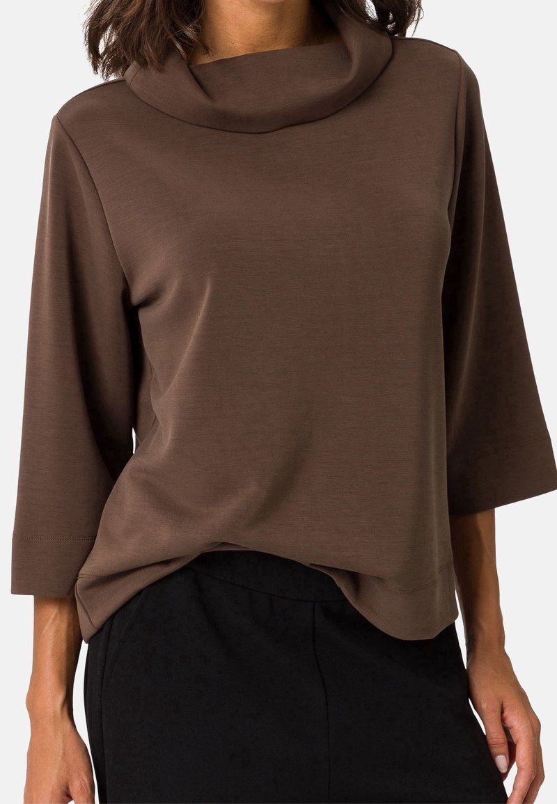 Femme portant un haut marron à manches trois-quarts avec un col roulé ample et un pantalon noir sur un fond uni.