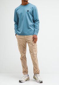 Helles blaues Langarmhemd mit einem kleinen schwarzen Logo auf der Brust, kombiniert mit beigen Cargo-Hosen mit Seitentaschen und mehrfarbigen Turnschuhen.