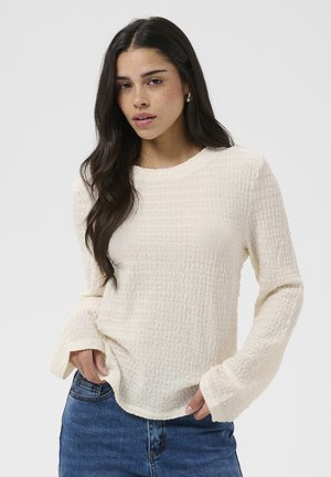 Jeune femme aux longs cheveux foncés porte un haut texturé à manches longues blanc cassé et un jean bleu, posant avec les mains près de la taille sur un fond uni.