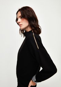 Schwarzes Sweatshirt mit hohem Kragen und goldenen Reißverschlussdetails an der Schulter. Gestreiftes Muster an den Bündchen. Weicher, strukturierten Stoff.