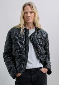 Chaqueta de plumas negra acolchada con textura brillante, cuello redondeado y cierre de cremallera. Llevada sobre una camiseta gris y pantalones negros.