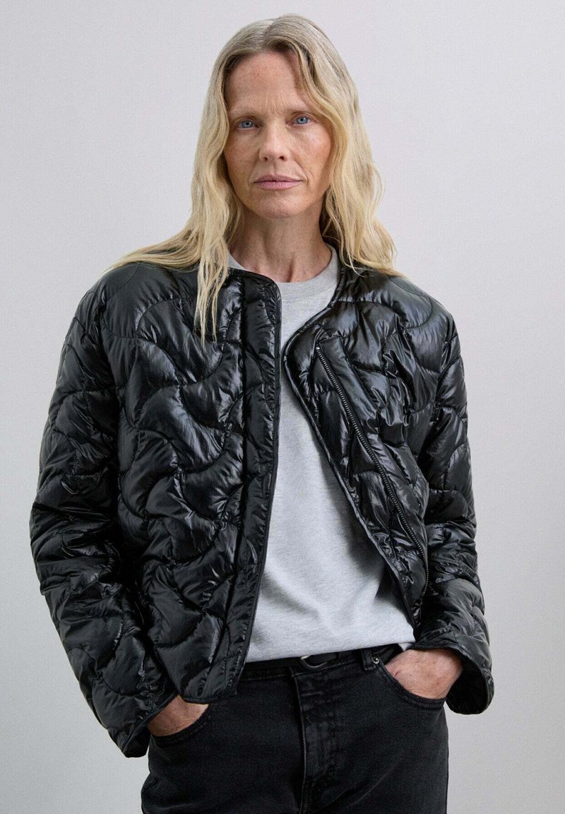 Chaqueta de plumas negra acolchada con textura brillante, cuello redondeado y cierre de cremallera. Llevada sobre una camiseta gris y pantalones negros.