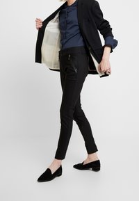Pantalon noir sur mesure avec une coupe slim, poches zippées et une texture lisse ; associé à un chemisier bleu marine et à un blazer noir.