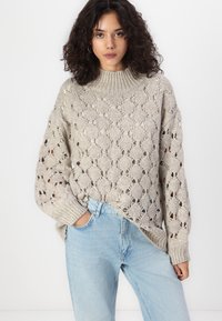 Chunky strikkegenser i lys beige med et teksturert, åpent strikkemønster. Har høy hals og ribbede mansjetter, kombinert med lyseblå jeans.