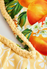 Prim-plan al unui material texturat cu imprimeu de fructe portocalii și frunze verzi, având o bordură împletită galbenă de-a lungul marginilor.