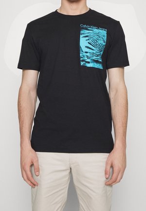 T-shirt en coton noir avec un motif graphique de feuilles de palmier turquoise sur la poitrine à gauche, avec le texte "Calvin Klein Jeans" au-dessus du design.