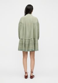 Γυναίκα με μακριά σκούρα μαλλιά που φοράει ένα φαρδύ, ανοιχτό πράσινο φόρεμα μέχρι το γόνατο με λεπτομέρειες από δαντέλα και καφέ δερμάτινα παπούτσια με κορδόνια, κοιτάζοντας προς τα πίσω σε απλό φόντο.