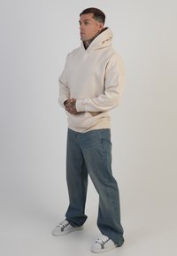 Gaiši brūna kapuces džemperis ar brīvu piegriezumu, kanguru kabatu un ribinātu piedurkņu acabumu, kopā ar gaiši ziliem džinsiem un baltām čizmām ar pelēkiem akcentiem.