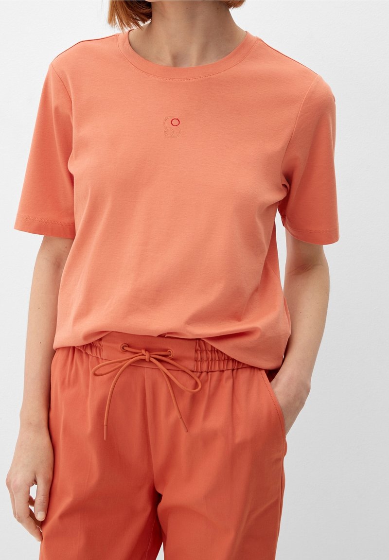 Orange bomulds t-shirt med korte ærmer og rund halsudskæring, som har et lille broderet design i kontrastfarver på brystet.