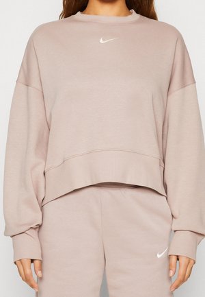 Sweat-shirt Nike beige oversize et pantalon de survêtement assorti avec logos Nike swoosh blancs sur la poitrine et la cuisse, porté par une personne.