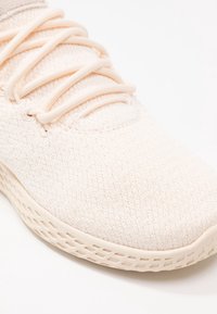 Ljust beige stickad sneaker med matchande snören och en texturerad sula på vit bakgrund.