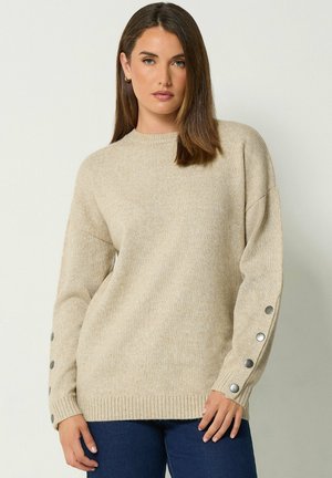 Mujer con un suéter de punto beige con botones plateados en las mangas y vaqueros azul oscuro, de pie frente a un fondo claro y liso.
