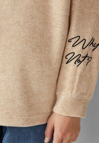 Jasnobeżowy sweter o miękkiej fakturze i luźnym kroju, z czarnym haftowanym napisem "Why Not?" na lewym rękawie.