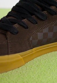 Baskets en daim marron avec un motif à carreaux sur le côté, lacets noirs, semelle en caoutchouc et un bord jaune vif près de la base.