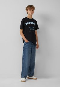 Schwarzes Baumwoll-T-Shirt mit blauer Schrift, kombiniert mit lockeren, hellblauen Jeans und weißen Sneakern mit beigen Akzenten.