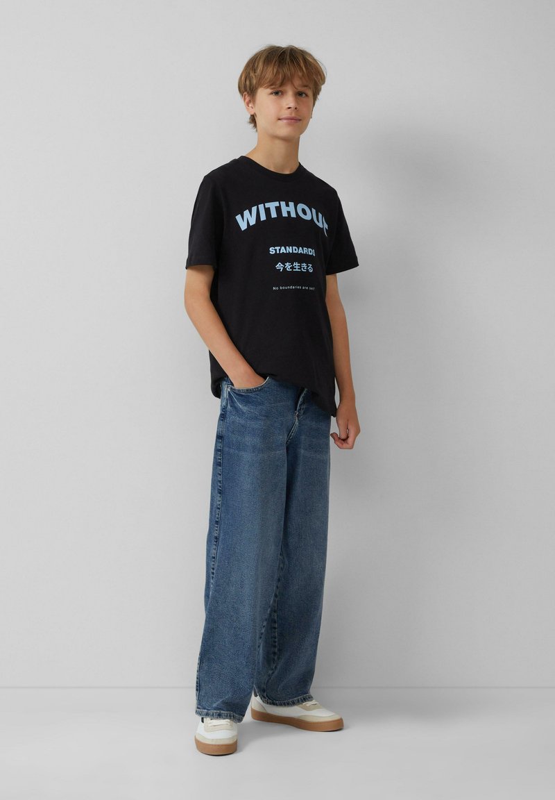 Schwarzes Baumwoll-T-Shirt mit blauer Schrift, kombiniert mit lockeren, hellblauen Jeans und weißen Sneakern mit beigen Akzenten.
