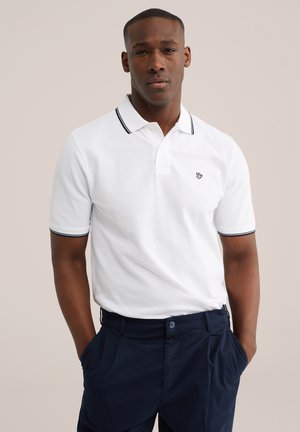 Mann trägt weißes Poloshirt mit schwarzer Einfassung und marineblaue Hose, steht mit den Händen in den Taschen vor einfarbigem Hintergrund.
