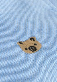 Material albastru cu o textură țesută, având o față mică de urs brodată, maro cu accente negre, poziționată ușor decentrată.