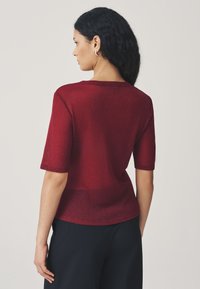 Rød kortærmet top med en glitrende tekstur, der har en afslappet pasform og rund halsudskæring. Bagfra ses blød drapering og glatte sømme.