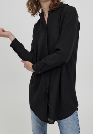 Osoba w czarnej, zapinanej na guziki koszuli z długim rękawem i jasnoniebieskich dżinsach, poprawiająca lewy rękaw prawą ręką na jednolitym, jasnym tle.