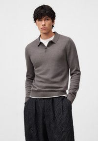 Pullover grigio a maglia con colletto e dettaglio dei bottoni, indossato sopra una maglietta bianca. Presenta maniche lunghe e orlo a coste, abbinato a pantaloni neri texturizzati.
