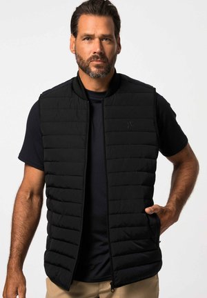 Chaleco aislante negro con patrón de acolchado vertical, cierre con cremallera y cuello acanalado, superpuesto a una camiseta negra. Cuenta con bolsillos laterales.