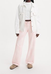 Femme portant un haut court blanc, une veste blanche à boutons, un jean large rose clair et des chaussures beiges à enfiler, se tenant sur un fond blanc.
