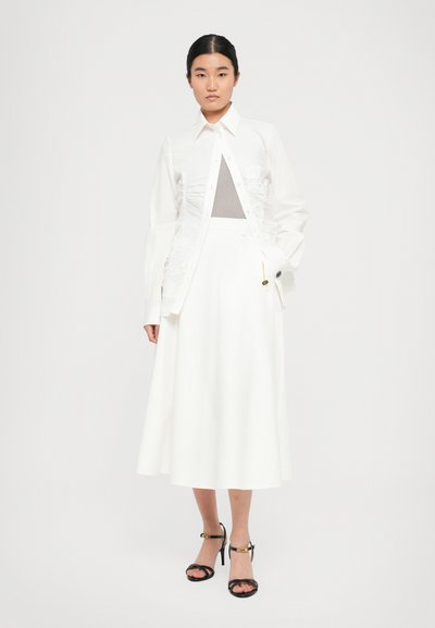 Woman with black hair wearing a white ruched button-up shirt, white midi skirt, gray top, and black high-heeled sandals standing against white background.