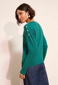 Zielony sweter z dzianiny z prążkowanym dołem, ozdobiony dekoracyjnymi złotymi guzikami na ramieniu. Ubrany z ciemnoniebieskimi spodniami z szerokimi nogawkami.