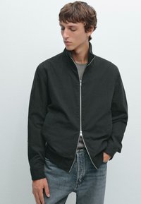 Black zip-up jacket with a stand collar, made of smooth fabric, paired with light blue denim jeans. Minimalistic design, no visible patterns.
