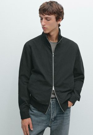 Svart zip-up-jacka med ståkrage, tillverkad av slät tyg, tillsammans med ljusblå jeans. Minimalistisk design, inga synliga mönster.