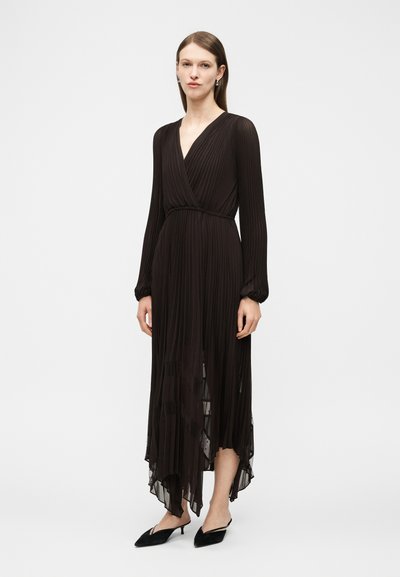 Γυναίκα που φοράει μακρυμάνικο μαύρο πλισέ φόρεμα μήκους μέχρι τη μέση της γάμπας με ασύμετρο τελείωμα και μαύρα μυτερά πέδιλα με λουράκι στο πίσω μέρος.