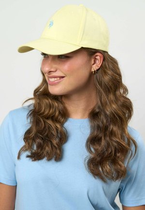 Tânără cu păr lung, creț și castaniu, purtând o șapcă galbenă de baseball și un tricou albastru deschis, zâmbind și uitându-se în lateral.