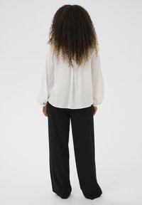 Personne aux cheveux longs et bouclés portant un chemisier blanc à manches longues et un pantalon noir ample, debout de dos sur un fond uni.