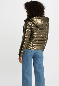 Chaqueta acolchada metálica verde oliva con capucha, que presenta secciones acolchadas horizontales y un logo en la manga izquierda. Combinada con unos vaqueros de pierna ancha en azul.