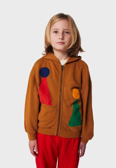 Felpa con zip marrone caratterizzata da vivaci patch geometrici in blu, rosso, verde e giallo. Tessuto morbido con una vestibilità rilassata, abbinata a pantaloni rossi.