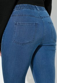 Sininen farkkutakki, jossa on kapea muotoilu, joustava vyötärö, kaksi takataskua ja sileä, hieman kiiltävä pinta.