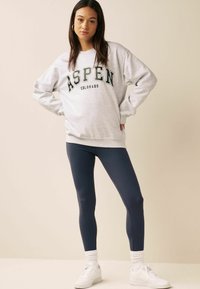 Siva dukserica s natpisom "ASPEN COLORADO", u kombinaciji s tamnoplave tajice i bijele tenisice. Ima opušteni kroj i rebraste manžete.