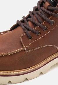 Stivale in pelle marrone con punta rotonda, lacci marroni e occhielli in ottone. Cuciture a contrasto e suola in gomma con un accento bordeaux.