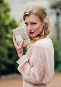 Blondýnka v světle růžové blůze drží béžovou peněženku blízko obličeje a s důvěrou se dívá na kameru venku.
