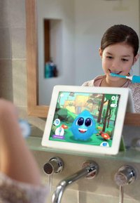 Ein Tablet mit einem hellen Rahmen zeigt eine blaue animierte Figur, die auf einer Glasoberfläche in einem Badezimmer platziert ist. Ein Kind putzt sich die Zähne.
