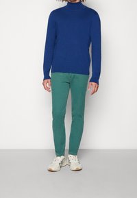 Niebieski sweter w golf z długimi rękawami, w zestawie z zielonymi spodniami o slim fit i jasnymi trampkami. Gładki materiał bez wzorów.