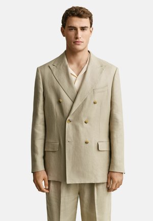 Jonge man die een beige dubbelknopaans pak draagt met tan knopen en een licht overhemd tegen een effen witte achtergrond.