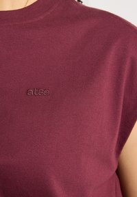 Gros plan sur une personne portant un débardeur bordeaux avec un petit texte brodé "ates" près de la clavicule.